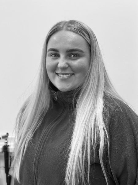 A woman with long, straight hair is smiling at the camera. She is wearing a zip-up jumper. The photo is in black and white, and the background is plain.