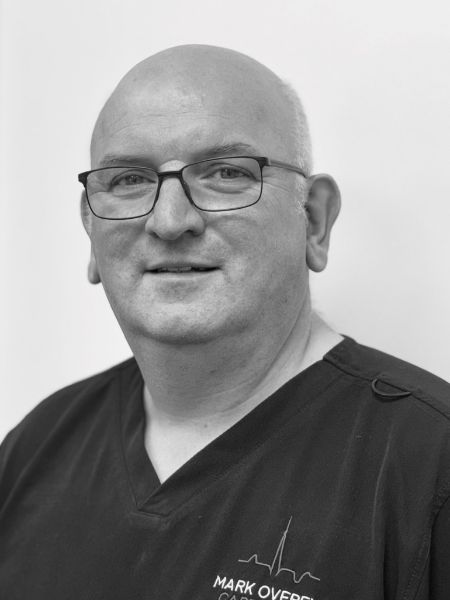A man with glasses and a shaved head, wearing a dark scrub shirt with the name Mark Over and a logo on it, is standing in front of a plain background.