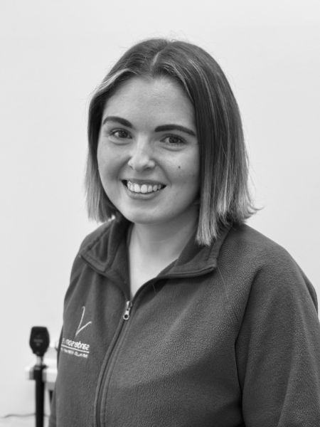 A woman with hair down to her shoulders and a zip-up jacket is smiling at the camera in a black-and-white photo. The background is blurry.