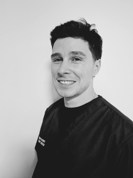 A person with short dark hair, dressed in medical scrubs, is standing in front of a plain white background and smiling at the camera. The photo is in black and white.