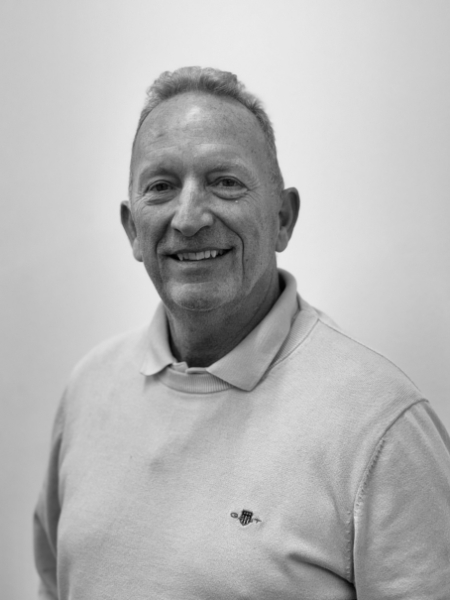 A black and white photo shows an older man with short hair, smiling at the camera. He is wearing a light-coloured jumper with a collar and is standing in front of a plain background.