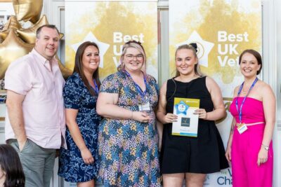 Five people are standing inside in front of Best UK Vets banners. One woman is holding a 2025 award and a certificate. They are all smiling and wearing event lanyards from Sanderson Vet.