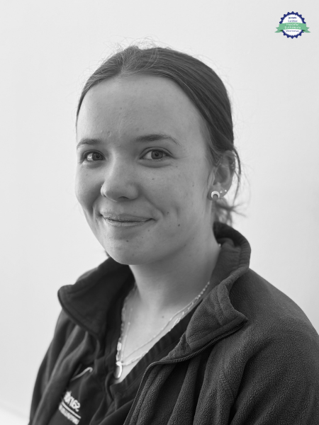 A young woman with straight hair tied back is wearing a zip-up jacket and a necklace. She faces the camera with a small smile. The background is plain, and there is a small logo in the top right corner.