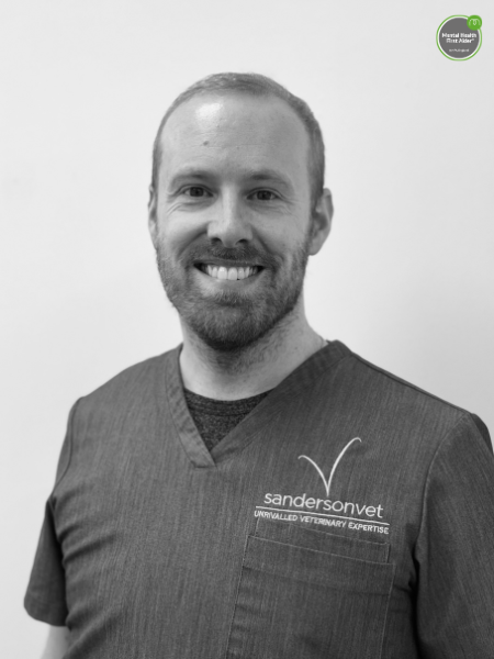 A man in scrubs with a sandersonvet logo is standing in front of a plain background, smiling at the camera.