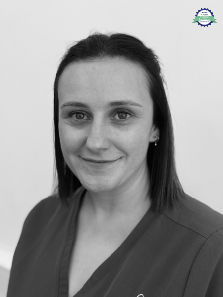 A woman with straight, dark hair to her shoulders wears a V-neck top and looks at the camera with a small smile. There is a small round certificate badge in the top right corner.