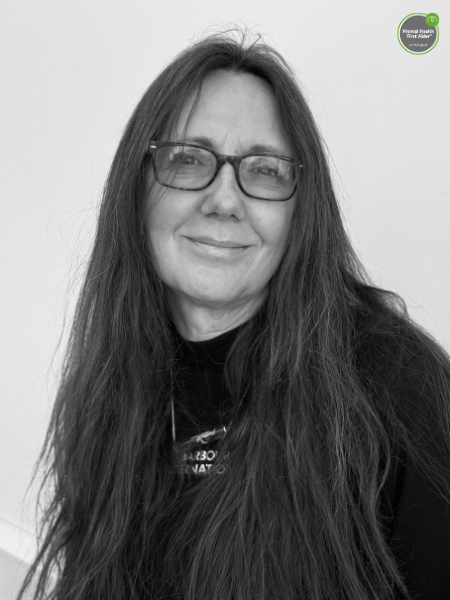 A woman with long dark hair and glasses is gently smiling at the camera. She is standing in front of a plain background.