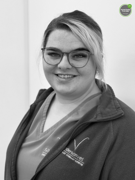 A woman with glasses and light-coloured hair tied back, wearing a fleece jacket and a scrub top, is standing in front of a white wall.