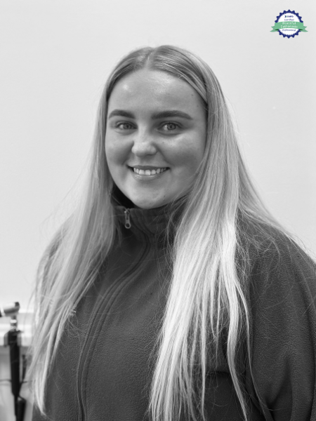 A young woman with long, straight hair and a zip-up jacket smiles at the camera in a black and white photo. There is a small blue and green emblem in the top right corner.