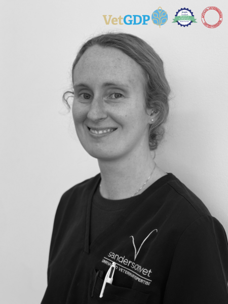 A woman wearing veterinary scrubs is smiling while standing in front of a plain background. There are logos related to veterinary work shown in the top right corner.