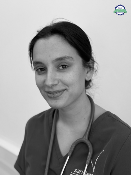 A black and white picture of a woman wearing medical scrubs and a stethoscope around her neck. She is looking at the camera with a small smile.