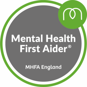 A round badge with “Mental Health First Aider” and “MHFA England” written on a grey background. The edge has green and white borders, and there is a leaf logo at the top right.
