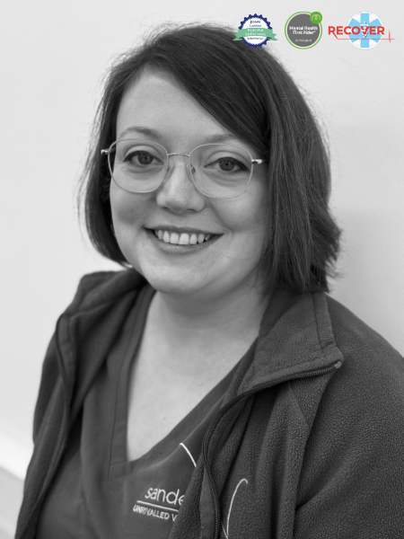 A black and white photo of a woman with glasses and short hair, smiling at the camera. She is wearing a jacket over her medical scrubs. Three certification logos are shown at the top.