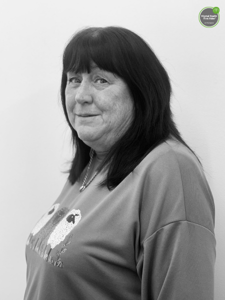 A woman with long dark hair and a fringe stands in front of a plain wall, wearing a shirt with a sheep on it. In the top right corner, there is a green badge that says Mental Health First Aider.