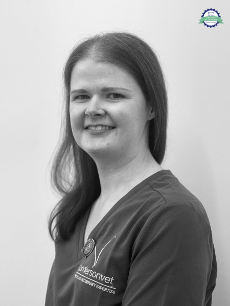 A woman with straight hair, wearing vet scrubs, stands in front of a plain background. She is turned a bit to the side and smiling.