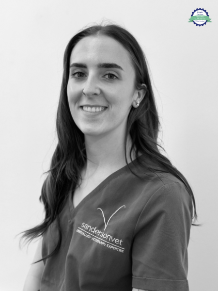 A black and white photo shows a woman in scrubs with a sandersonvet logo. She stands in front of a plain background and gives a small smile to the camera.