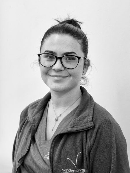 A woman with dark hair tied back is wearing glasses, a necklace, and a zip-up jacket over her scrubs. She is standing in front of a plain light-coloured background.