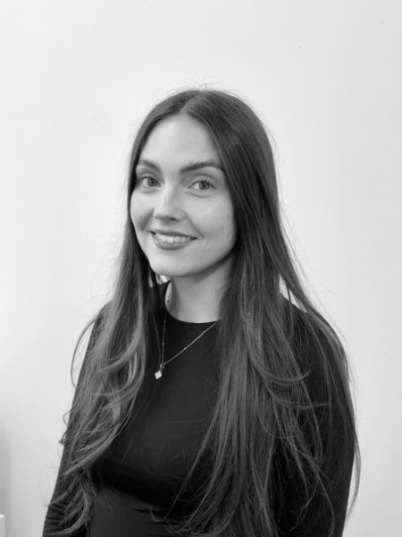 A woman with long straight hair is wearing a dark shirt and a necklace. She is smiling at the camera, and there is a plain light background behind her.