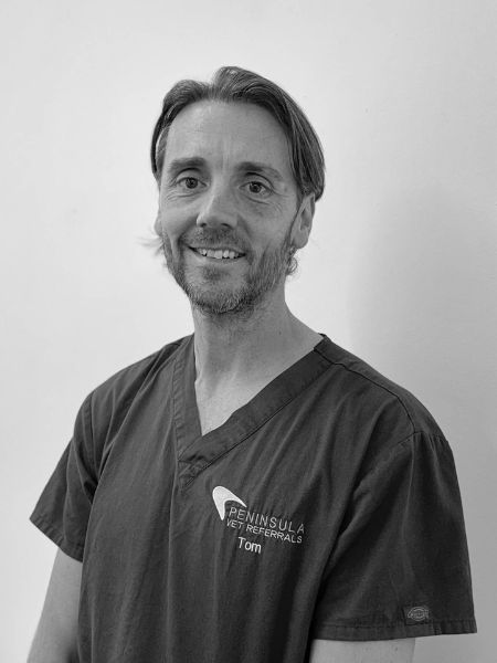 A man in scrubs with “Peninsula Vet Referrals” and the name “Tom” on his shirt is standing and smiling at the camera in front of a plain background.
