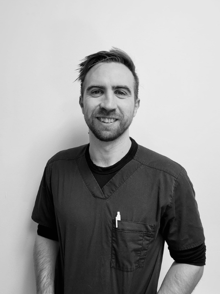 A man in scrubs with a pen in his chest pocket stands by a plain wall, smiling at the camera.