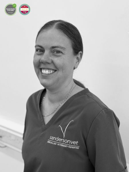 A woman wearing veterinary scrubs with a sandersonvet logo is smiling at the camera in a black and white photo. Two small award badges can be seen in the top left corner.