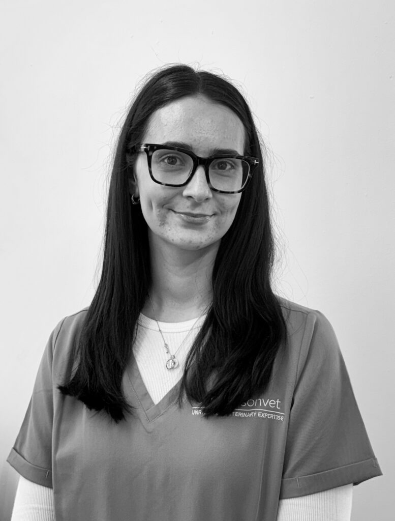 A woman with long dark hair and glasses is wearing a scrub top over a white vest. She is standing in front of a plain, light-coloured background.