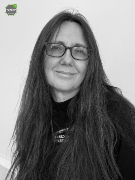 A woman with long dark hair and glasses gives a small smile whilst sitting in front of a plain light-coloured background.