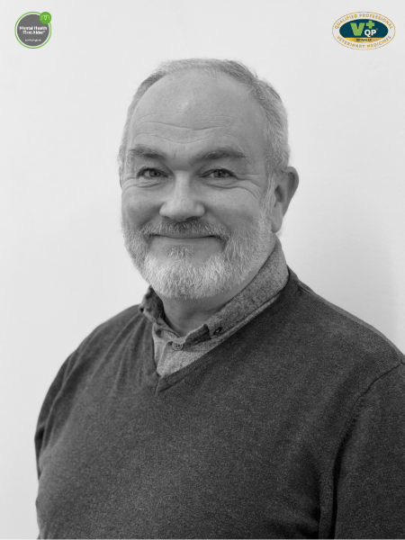 Black and white photo of an older man with a beard, smiling. He is wearing a collared shirt and a jumper. There are two logos in the corners of the picture.