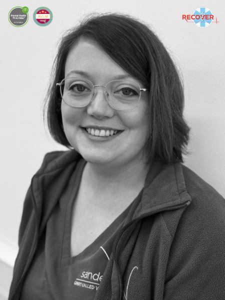 A black and white photo shows a woman with short hair and glasses, smiling at the camera. She is dressed in scrubs and a jacket. At the top corners, there are two round badges and a logo.