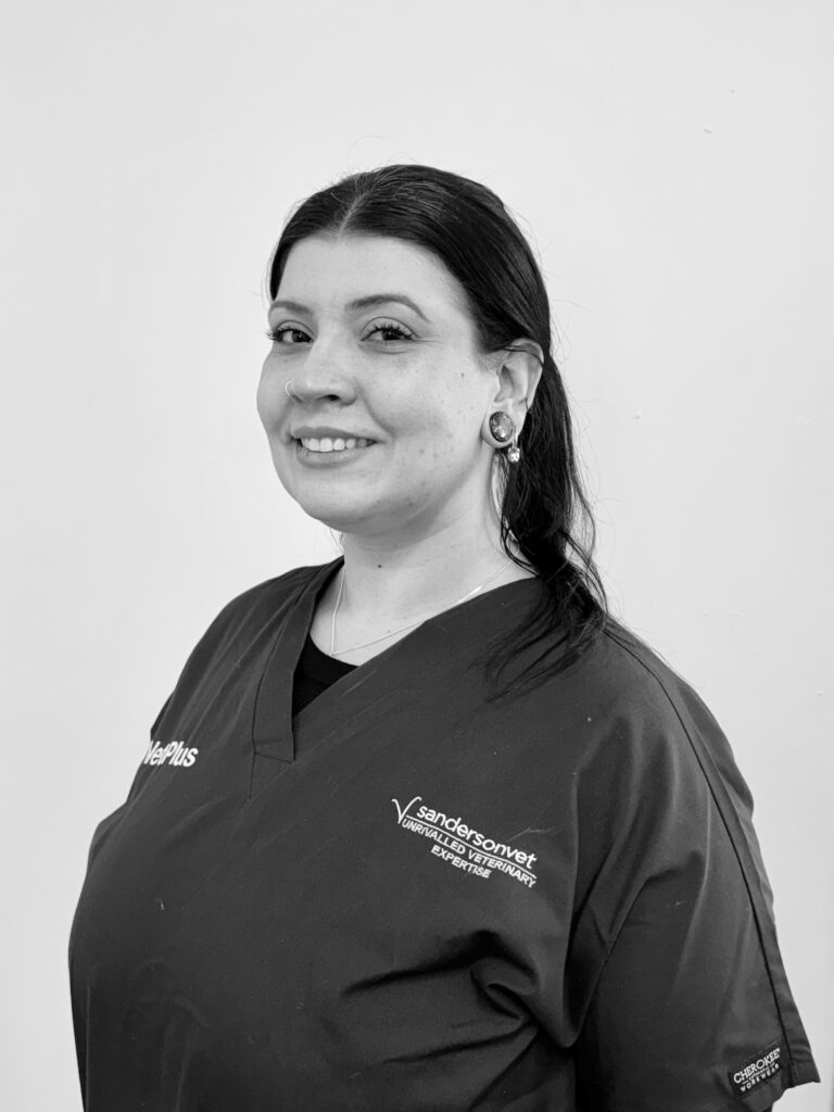 A woman wearing medical scrubs stands in front of a plain background and smiles. Her scrubs have the Sanders Onvet logo and a name tag on her chest.
