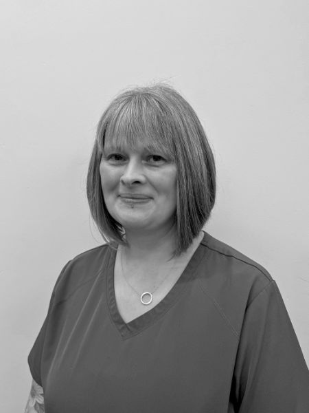 A woman with straight, shoulder-length hair is wearing a simple top and a necklace. She stands in front of a plain wall, looking at the camera with a calm face. The photo is in black and white.