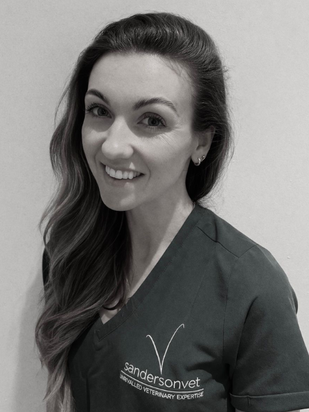 A woman with long hair is smiling at the camera. She is wearing a dark scrub top that has the sandersonvet logo and the words UNRIVALLED VETERINARY EXPERTISE on it.
