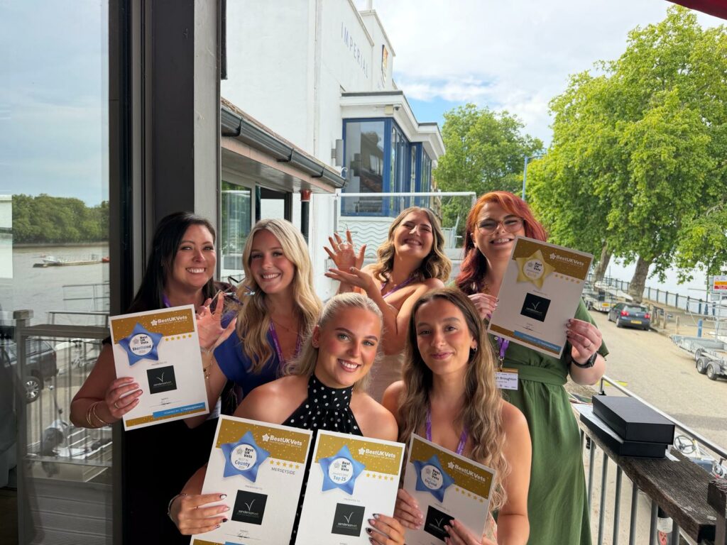 Six women are standing outside together, smiling happily and holding certificates and awards for winning the Sanderson Vet Best UK Vet award. There are trees, a river, and some buildings behind them.