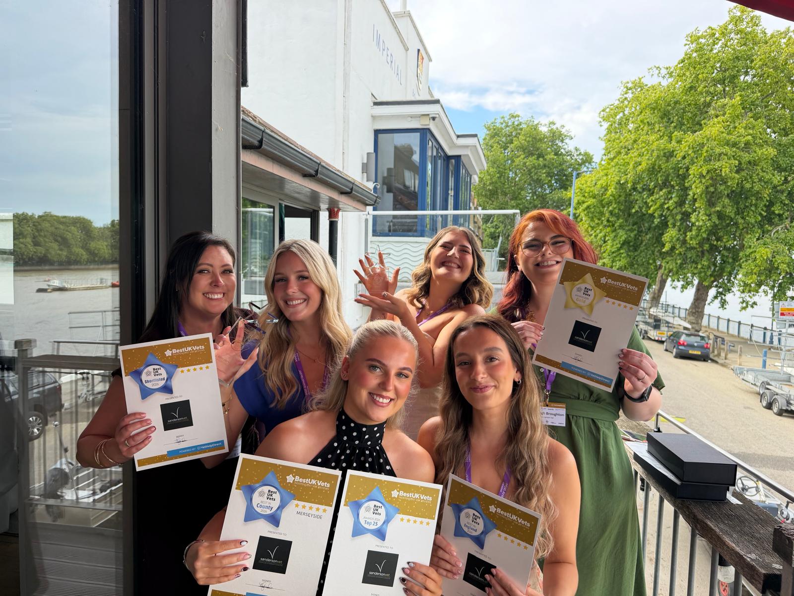 Six women are standing outside together, smiling happily and holding certificates and awards for winning the Sanderson Vet Best UK Vet award. There are trees, a river, and some buildings behind them.